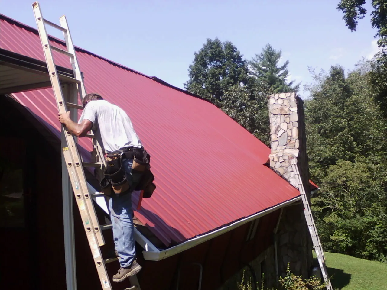 Roofer on a ladder beside a red metal roof and stone chimney — Soffit & Fascia Repair by Roofing Brothers in Bahama